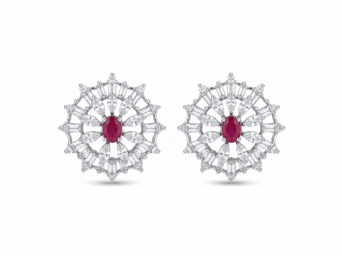 A pair of round silver-toned stud earrings featuring a central oval ruby gemstone surrounded by radiating baguette and round-cut clear stones, shown on a plain white background.
