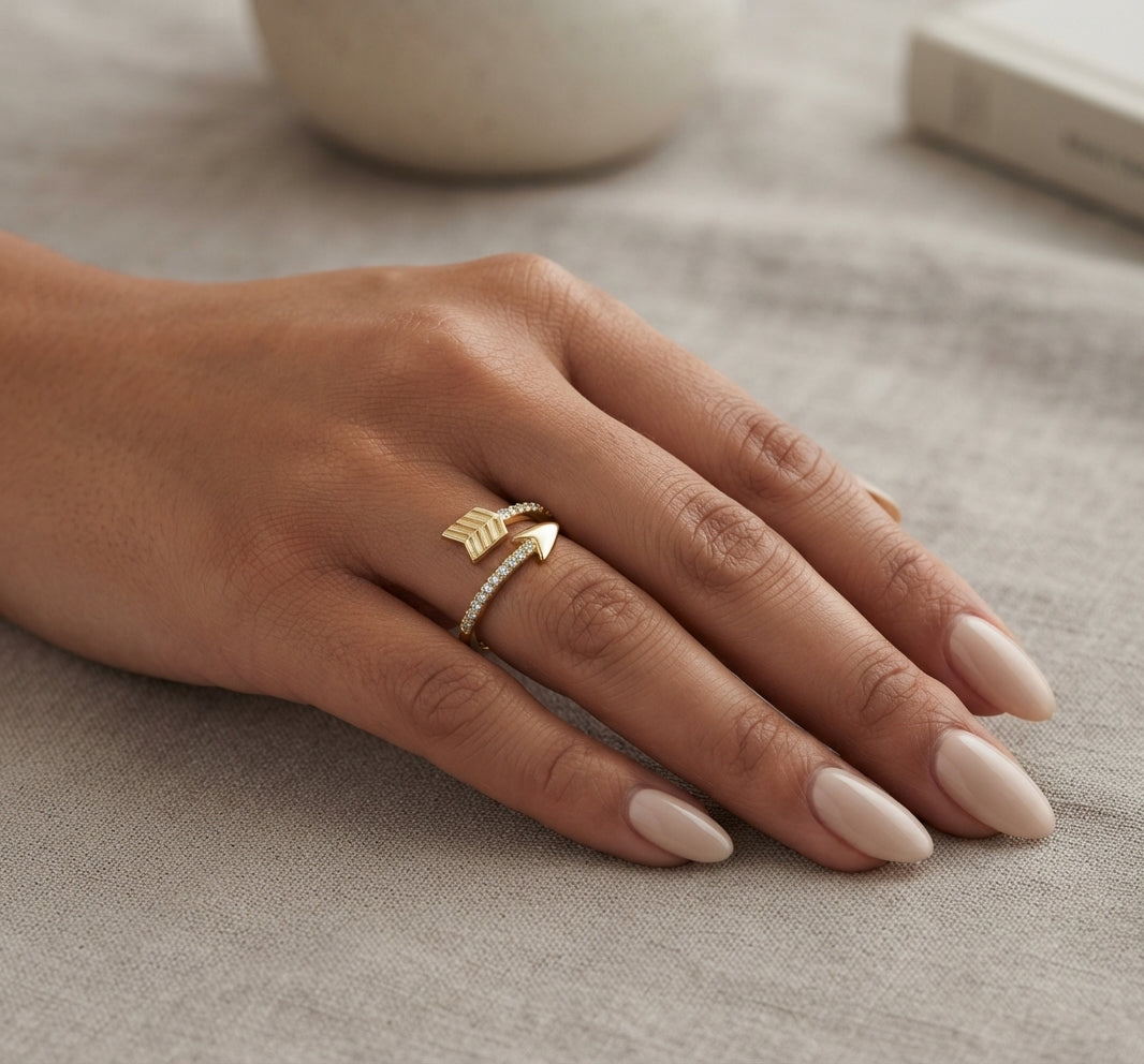 A close-up of a hand with long nude-pink manicured nails wearing two delicate rings: one gold arrow ring and one thin gold band with small clear stones, resting on a textured beige fabric surface.