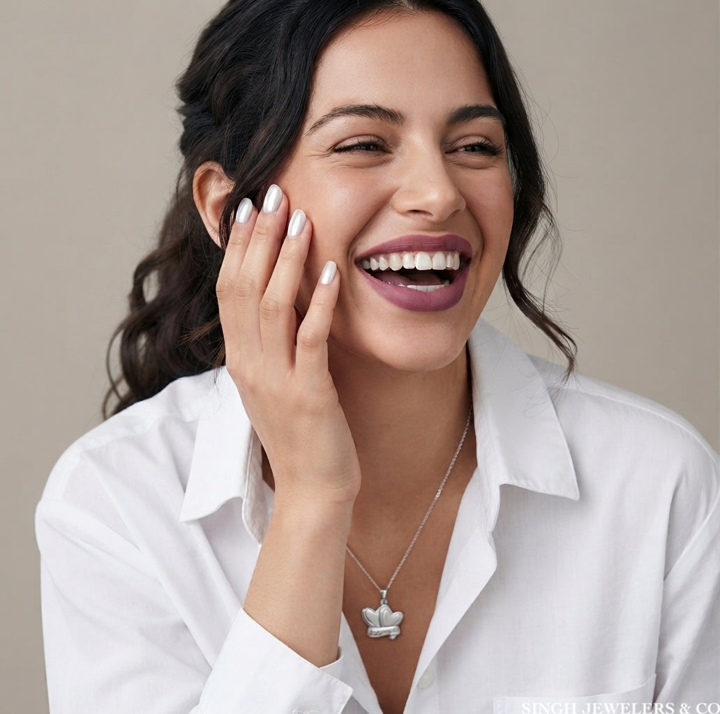 A woman in a white collared shirt smiling and touching her face, wearing a delicate silver necklace with a small crown pendant. The background is a solid, warm neutral beige.