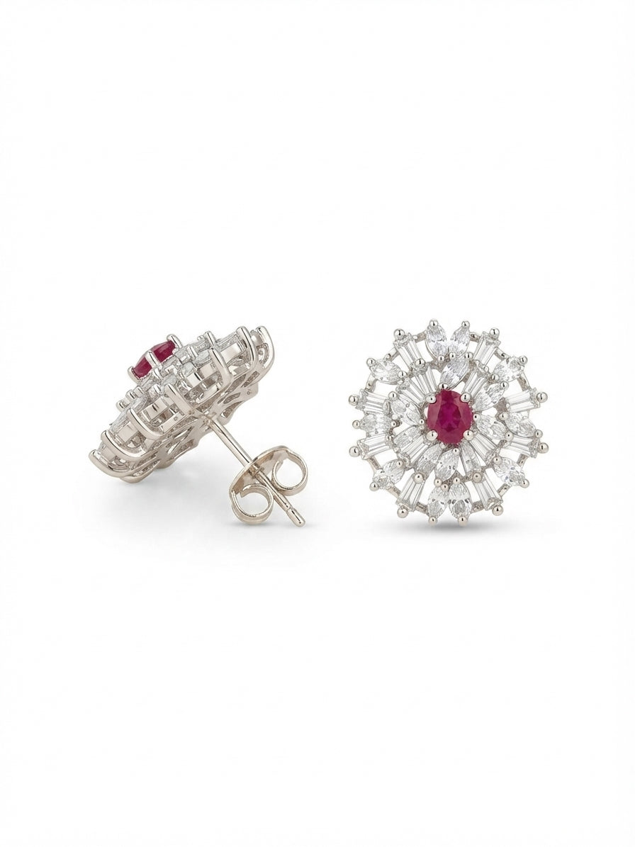 A pair of silver-toned stud earrings featuring a central round ruby red gemstone surrounded by a halo of small clear stones, shown on a plain white background with one earring facing forward and the other angled to show the post and back.