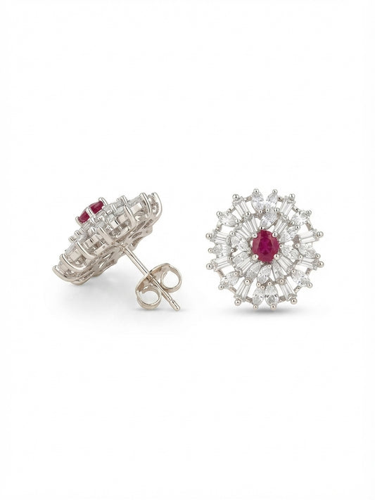 A pair of silver-toned stud earrings featuring a central round ruby red gemstone surrounded by a halo of small clear stones, shown on a plain white background with one earring facing forward and the other angled to show the post and back.